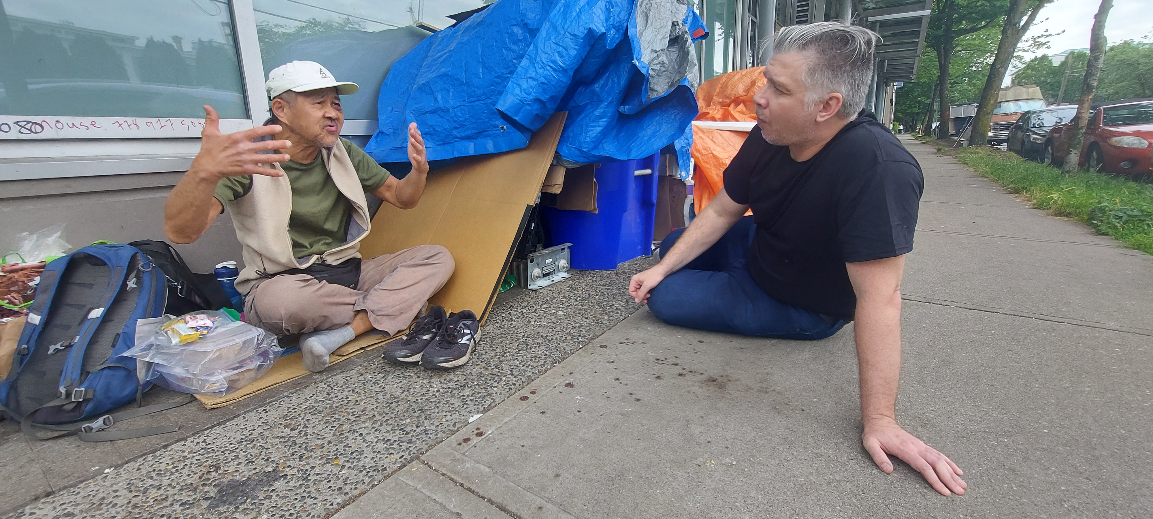 A field conversation on a sidewalk: one person seated beside a tarp encampment, another sitting opposite on the pavement, mid-exchange.